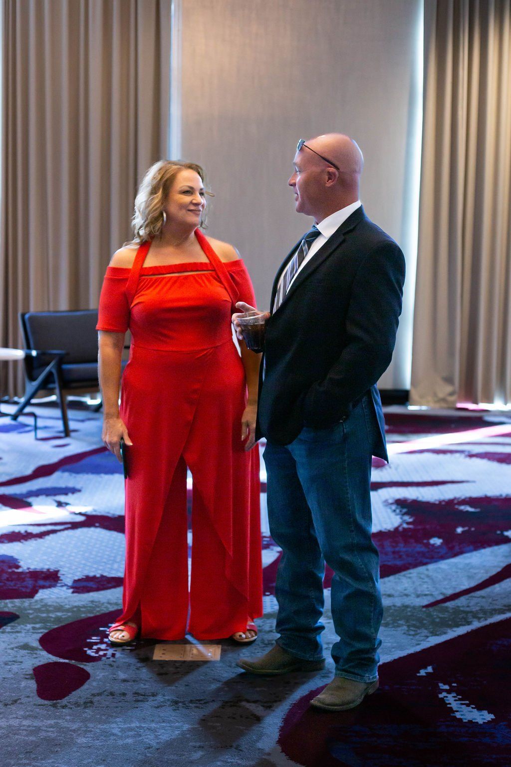 A person in a red jumpsuit talks to a person in a dark blazer and blue jeans in a room with floor-to-ceiling curtains.