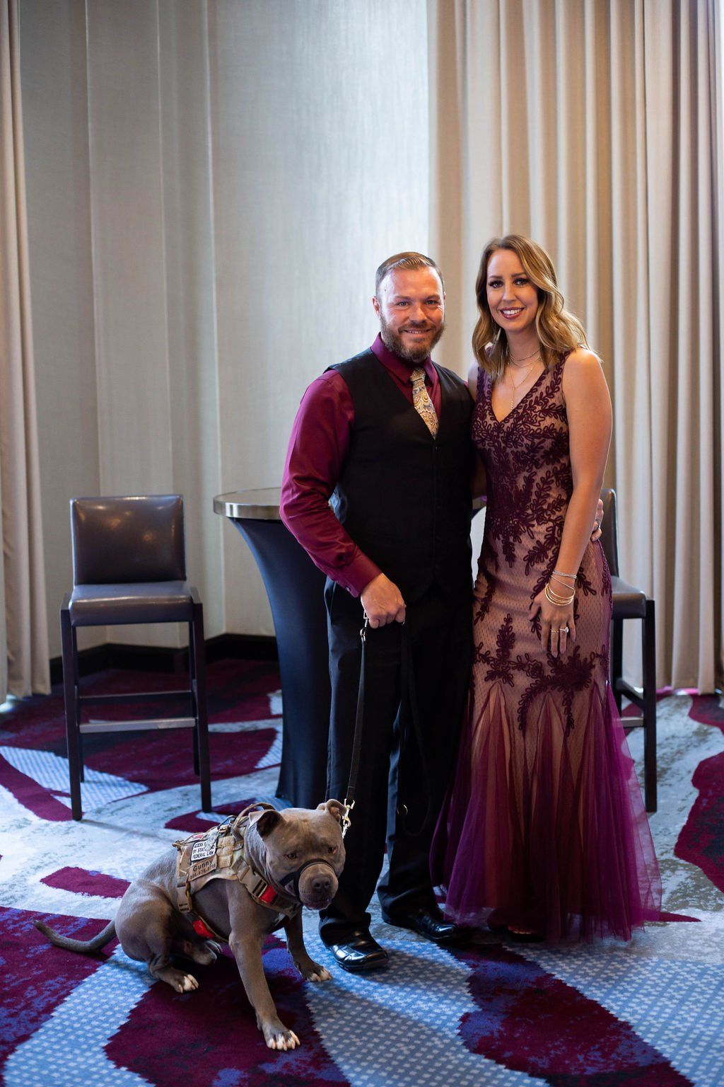 A man in a vest and a woman in a formal gown stand indoors with a dog wearing a harness on a patterned carpet.