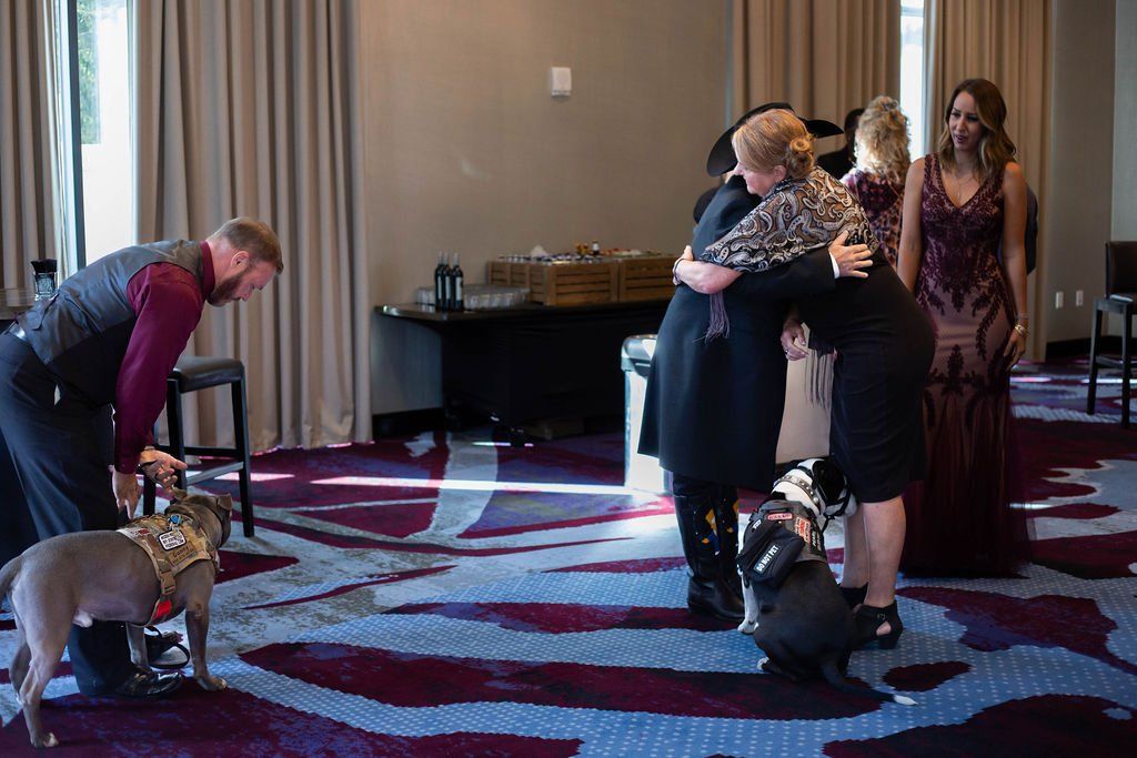 A man and three women are in a carpeted room with two service dogs. Two women embrace while another stands nearby.
