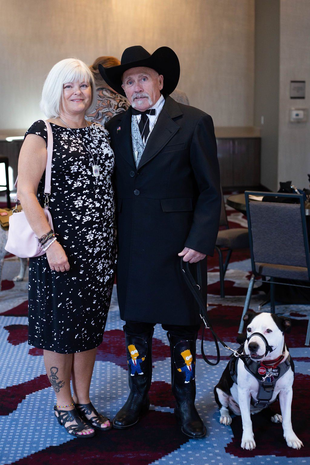A woman in a black patterned dress stands with a man in a black coat, cowboy hat, and cartoon-themed boots, with a dog.