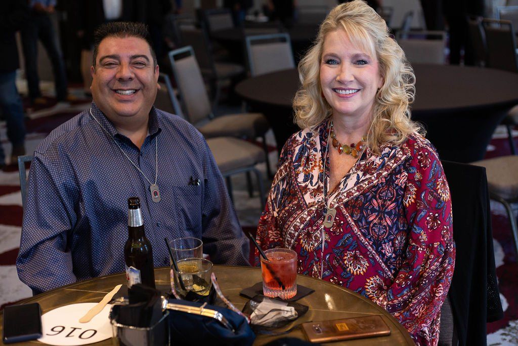 A smiling man and woman sit at a round table with drinks and a table number in a hotel ballroom setting.