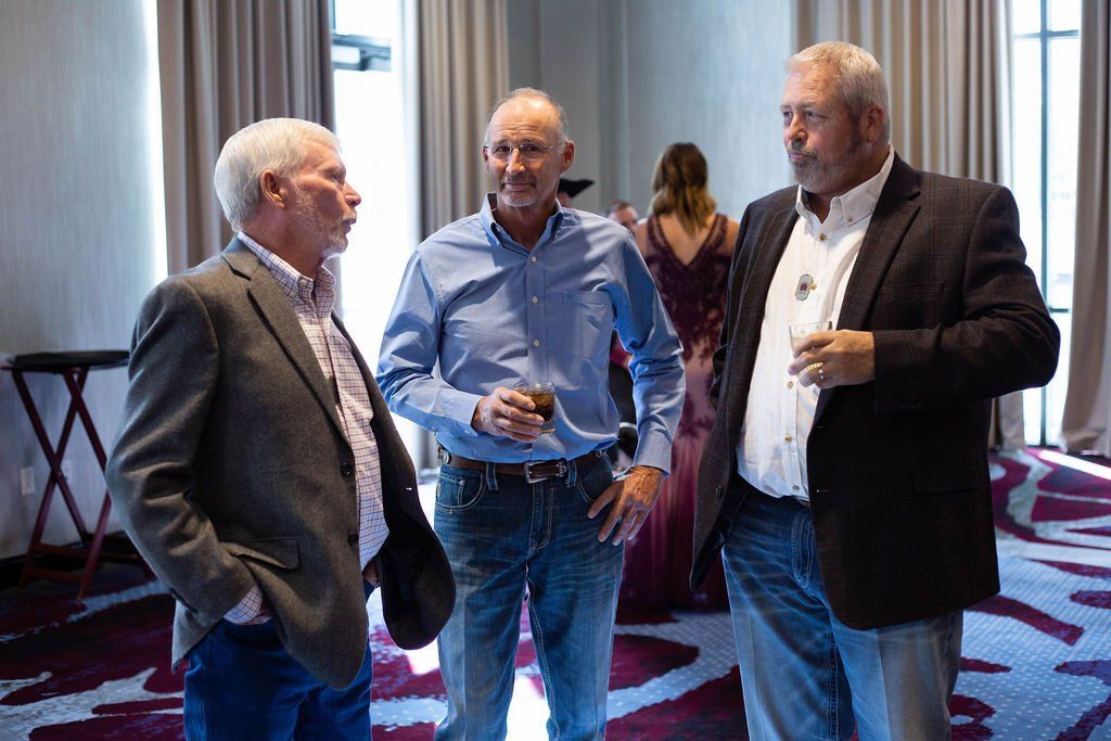 Three people in business-casual attire standing and talking in a hotel event space with patterned carpet.