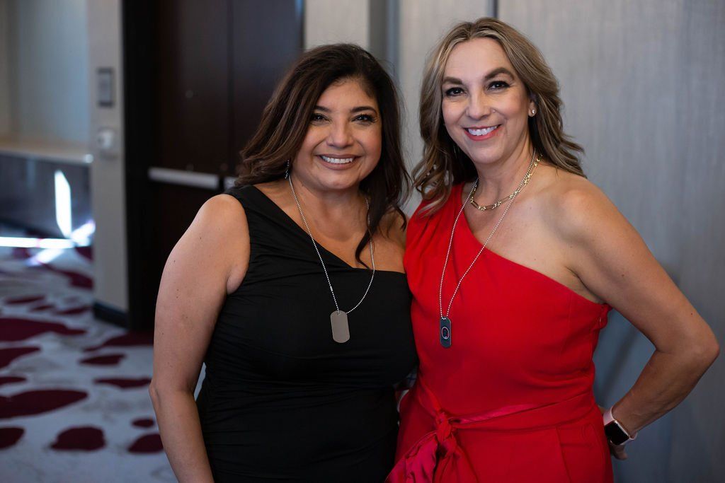 Two women smiling side-by-side, one in a black one-shoulder dress and the other in a red one-shoulder dress.