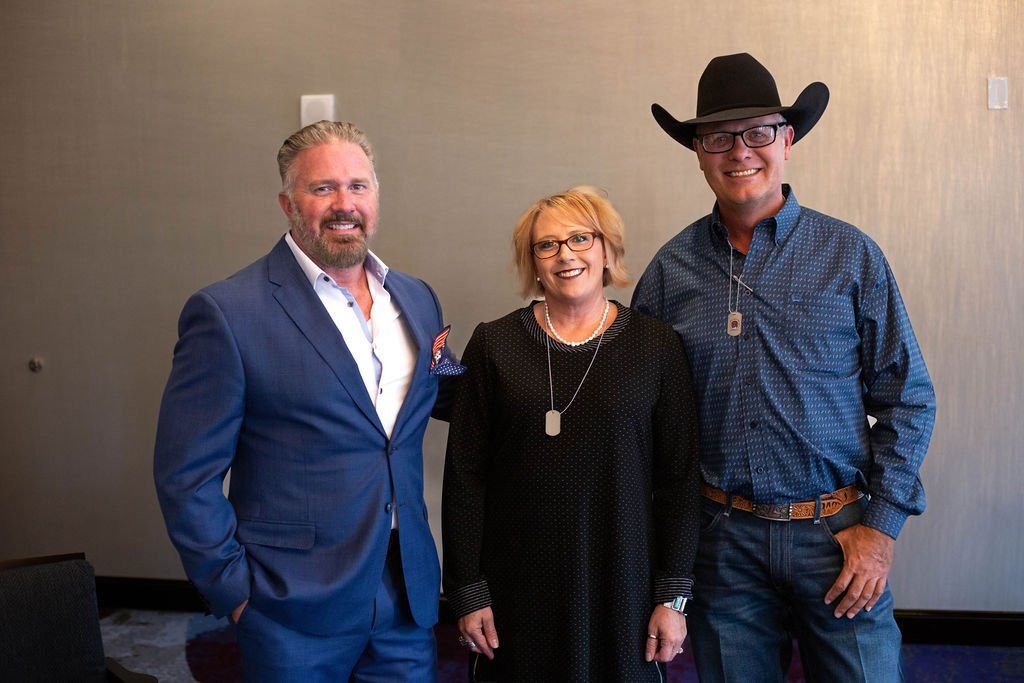 Three people posing for a photo indoors; a man in a blue suit, a person in a black dress, and a man in a cowboy hat.