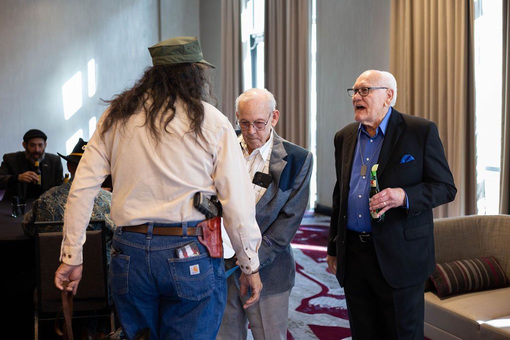 A person in a hat and jeans interacts with two men in suits during an indoor gathering.