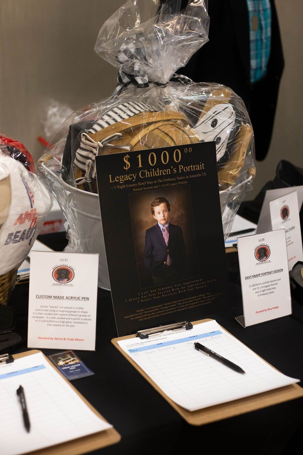 Silent auction table with a wrapped gift basket, a portrait voucher display, and two clipboards with pens on black cloth.