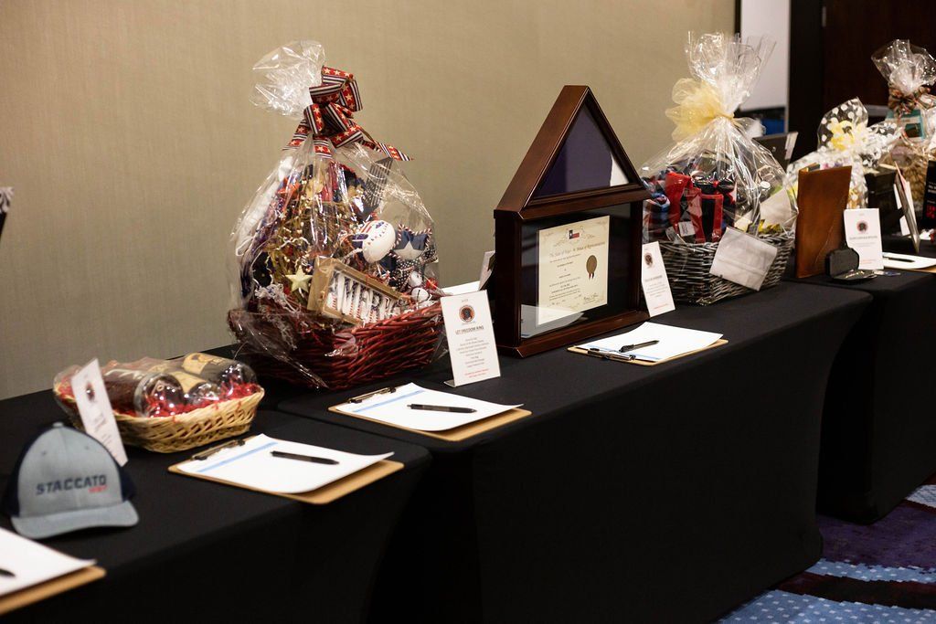 A row of silent auction baskets and items on tables covered with black tablecloths, including clipboards and a cap.