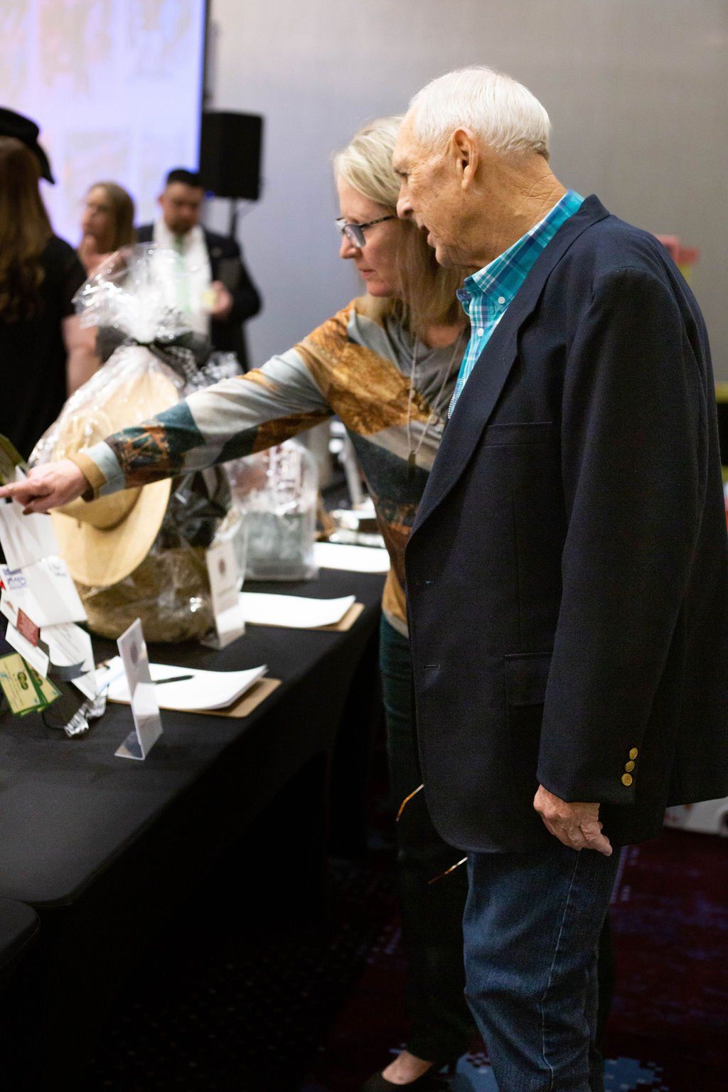 A person points toward a raffle basket on a table while an older companion looks on in an indoor event space.