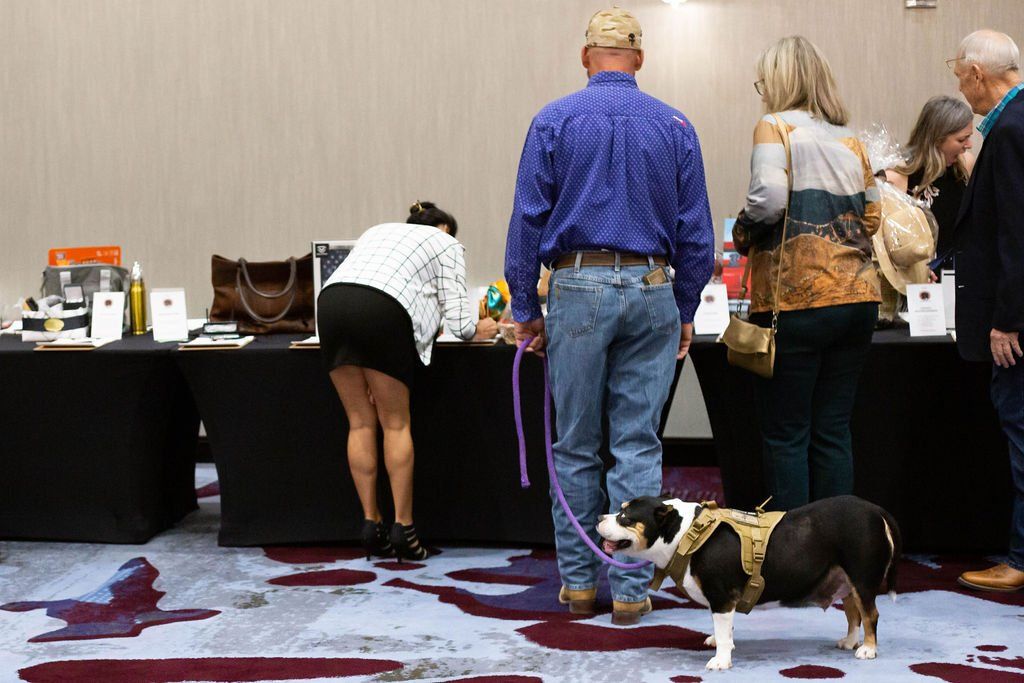 People gather around tables at an event, with a leashed dog standing near a man in a blue shirt.
