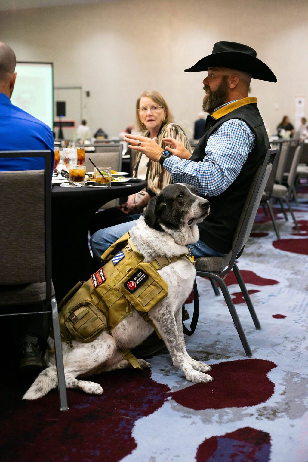 A service dog in a tan tactical vest sits next to a man wearing a cowboy hat at a table in a conference room.