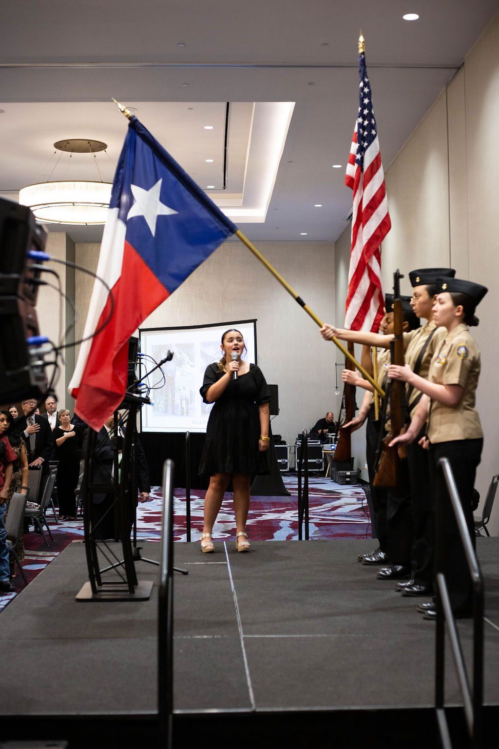 A person in a black dress speaks at a podium while a military color guard stands to the side holding U.S. and Texas flags.
