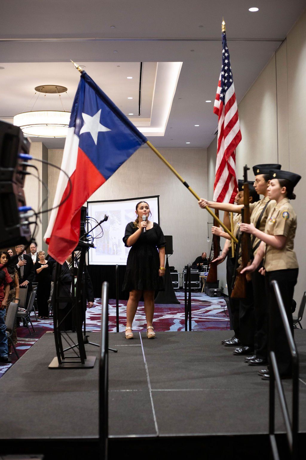 A woman speaks at a podium as a color guard holds the Texas and American flags during an indoor ceremony.
