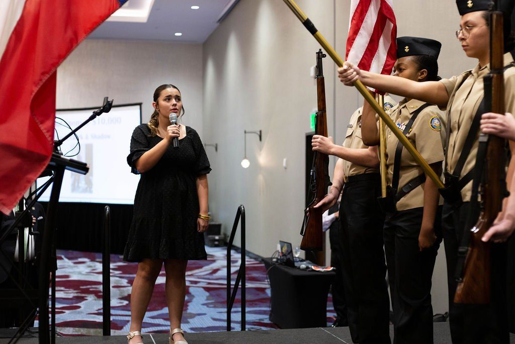 A woman speaks into a microphone on stage near a color guard holding the American flag and ceremonial rifles indoors.