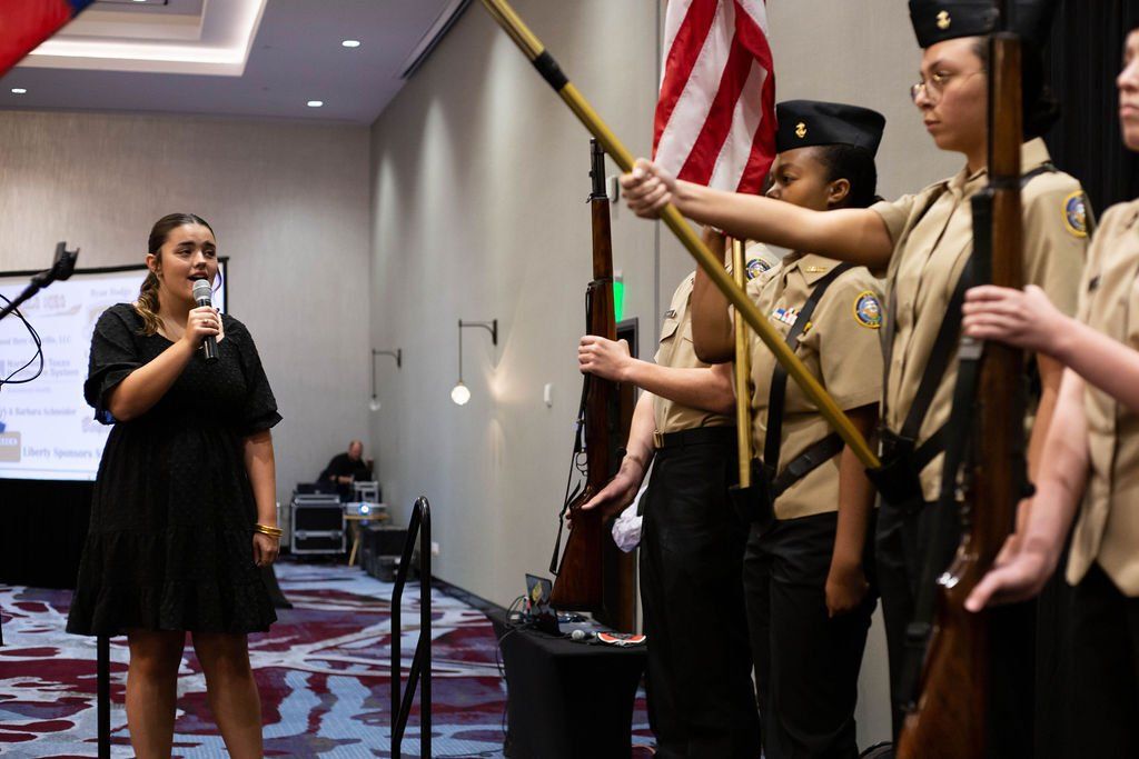A person in a black dress speaks into a microphone as a color guard in tan uniforms holds flags and rifles indoors.