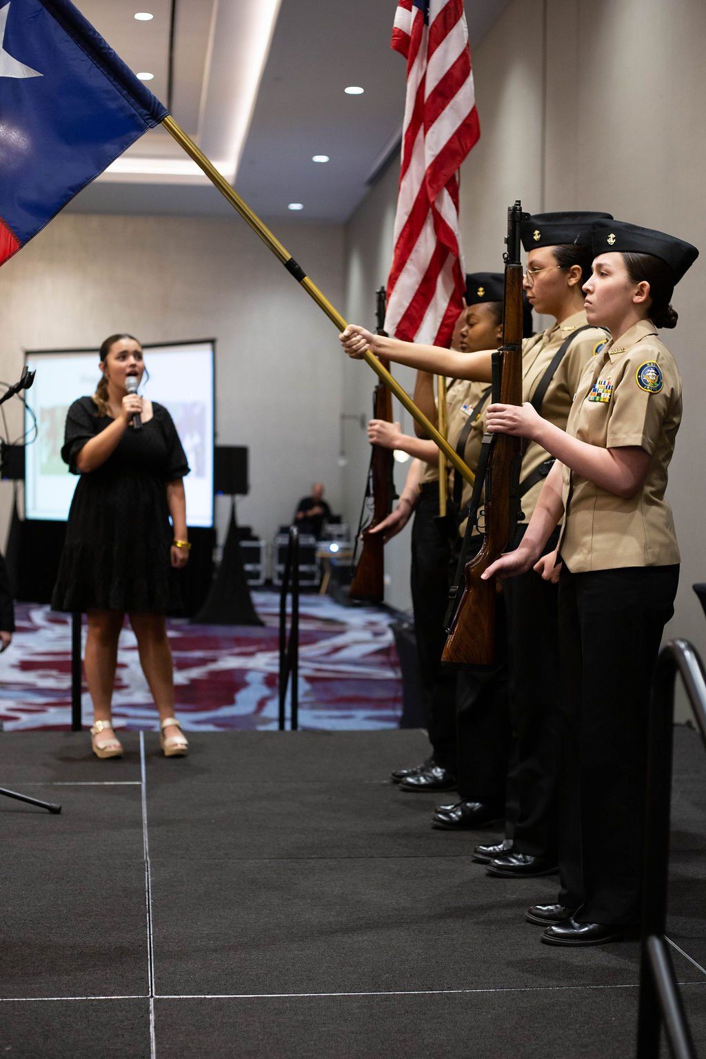 A person speaks into a microphone while a color guard stands in formation with US and Texas flags on an indoor stage.
