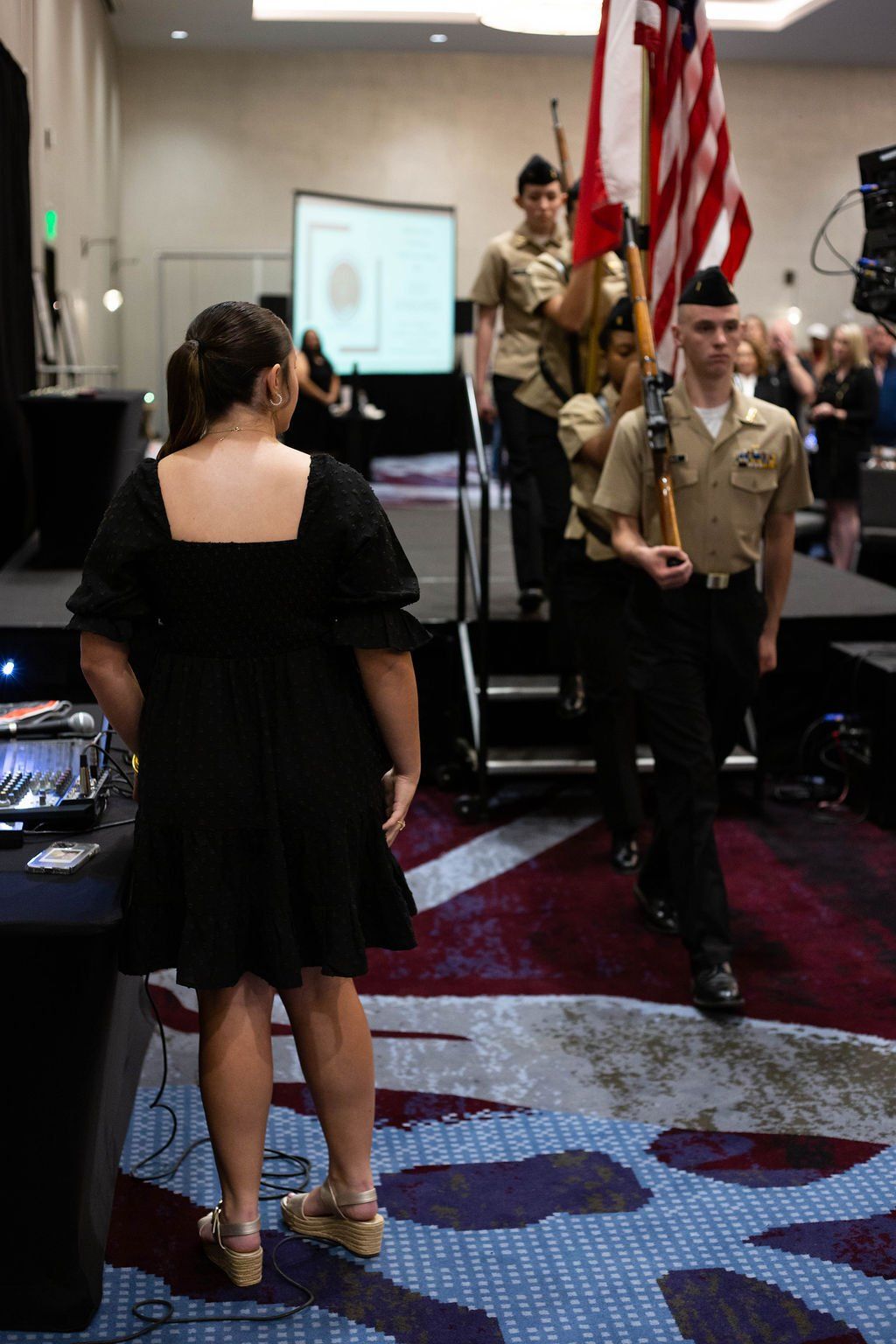 A woman in a black dress watches as a color guard in uniform carries the American flag during an indoor event.