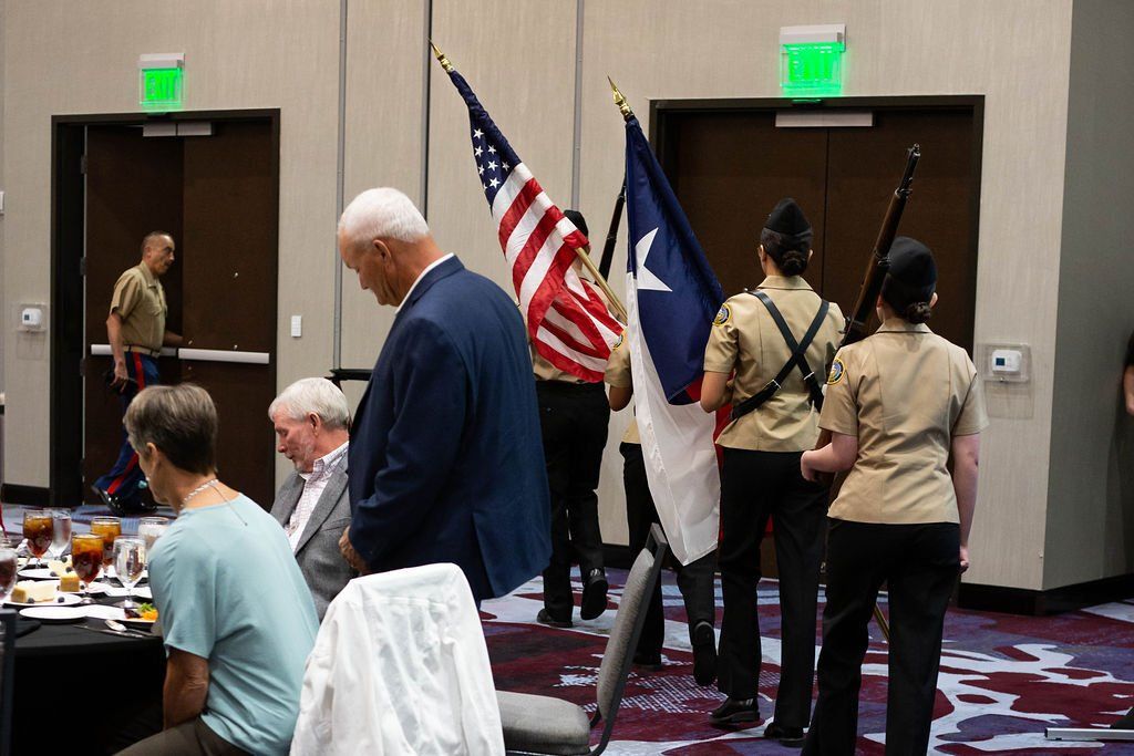 A color guard carrying US and Texas flags stands at the back of a room while people dine at tables in the foreground.