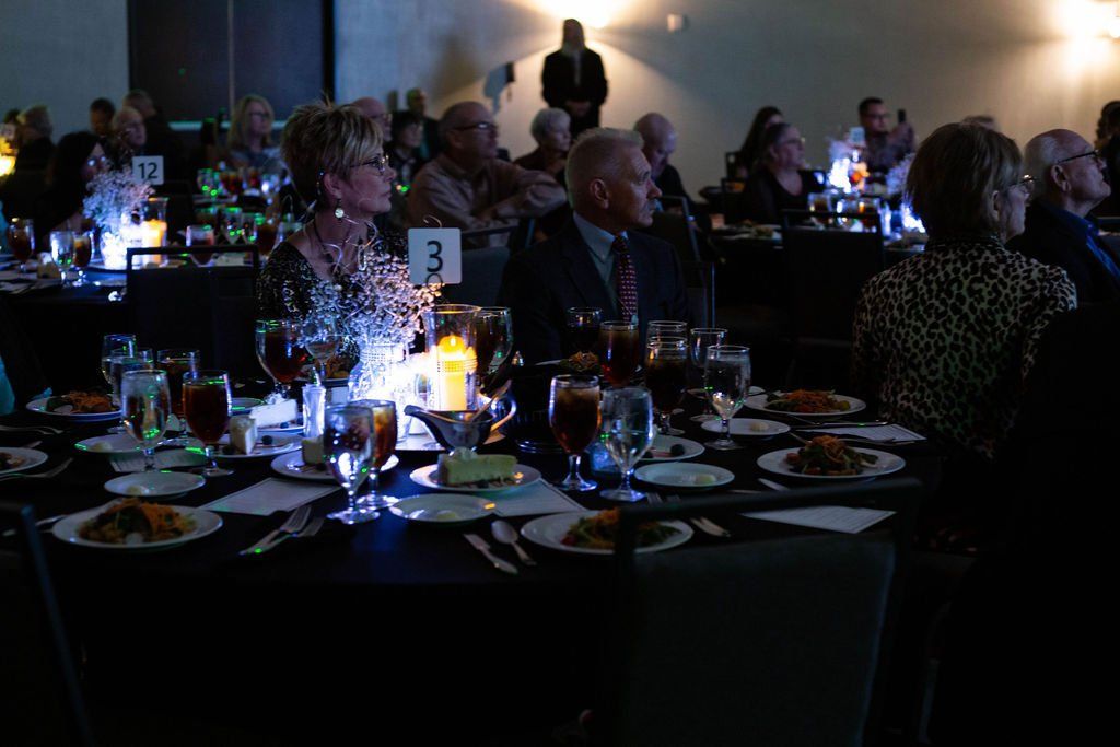 People sit at dinner tables in a dimly lit banquet hall, illuminated by glowing table centerpieces.
