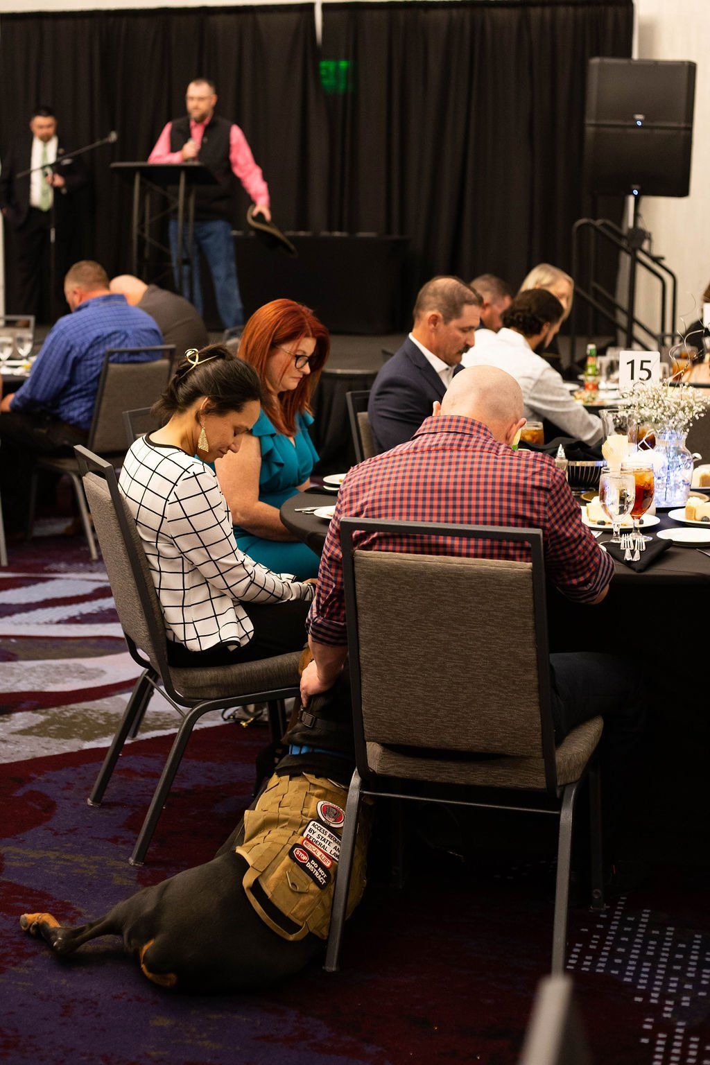 A service dog with a tan vest rests on the floor under a chair at a banquet table where guests are seated.