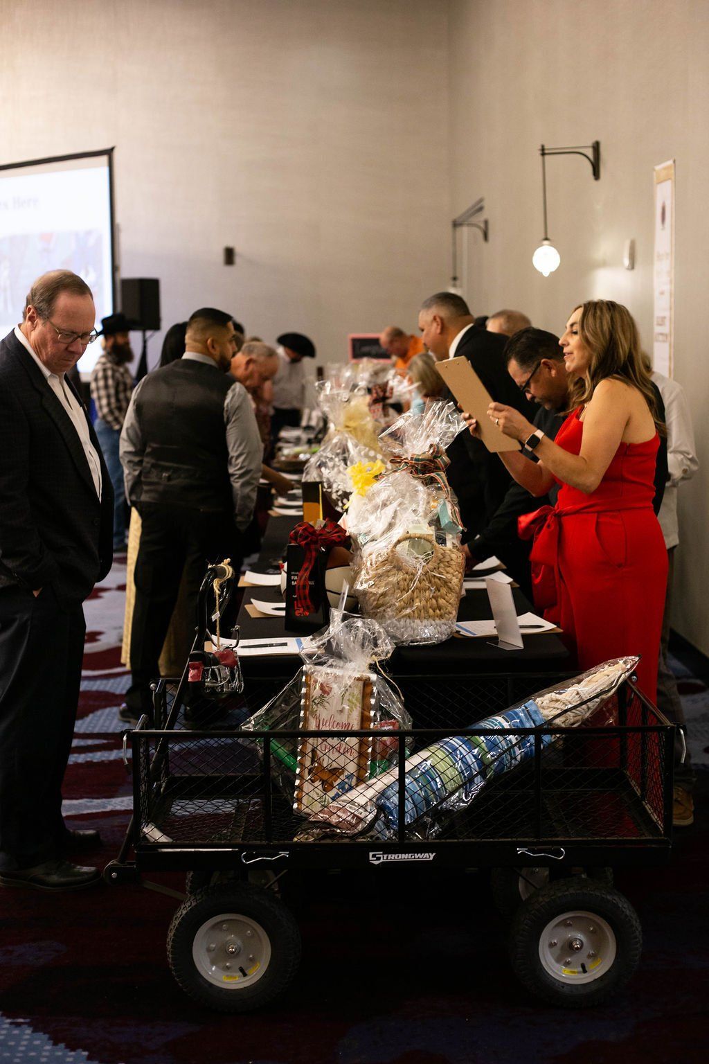 People browse a display of gift baskets arranged on a table next to a black utility cart in an indoor event space.