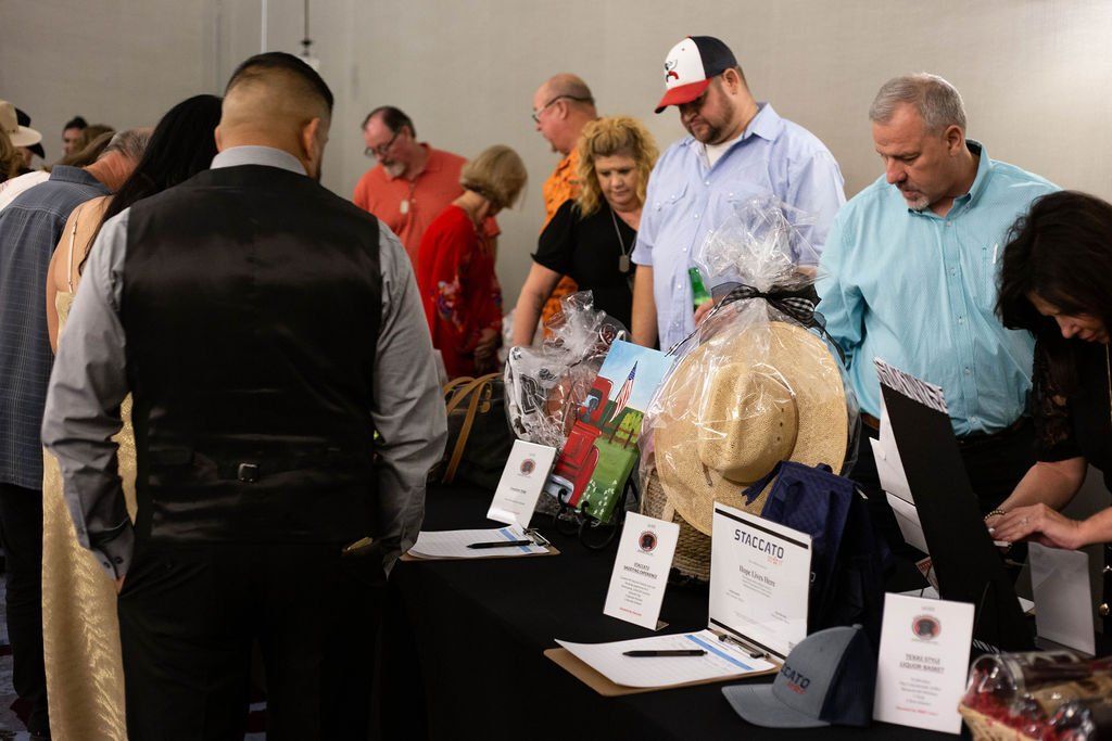 People browsing silent auction items displayed on a long table with bid sheets and gift baskets.