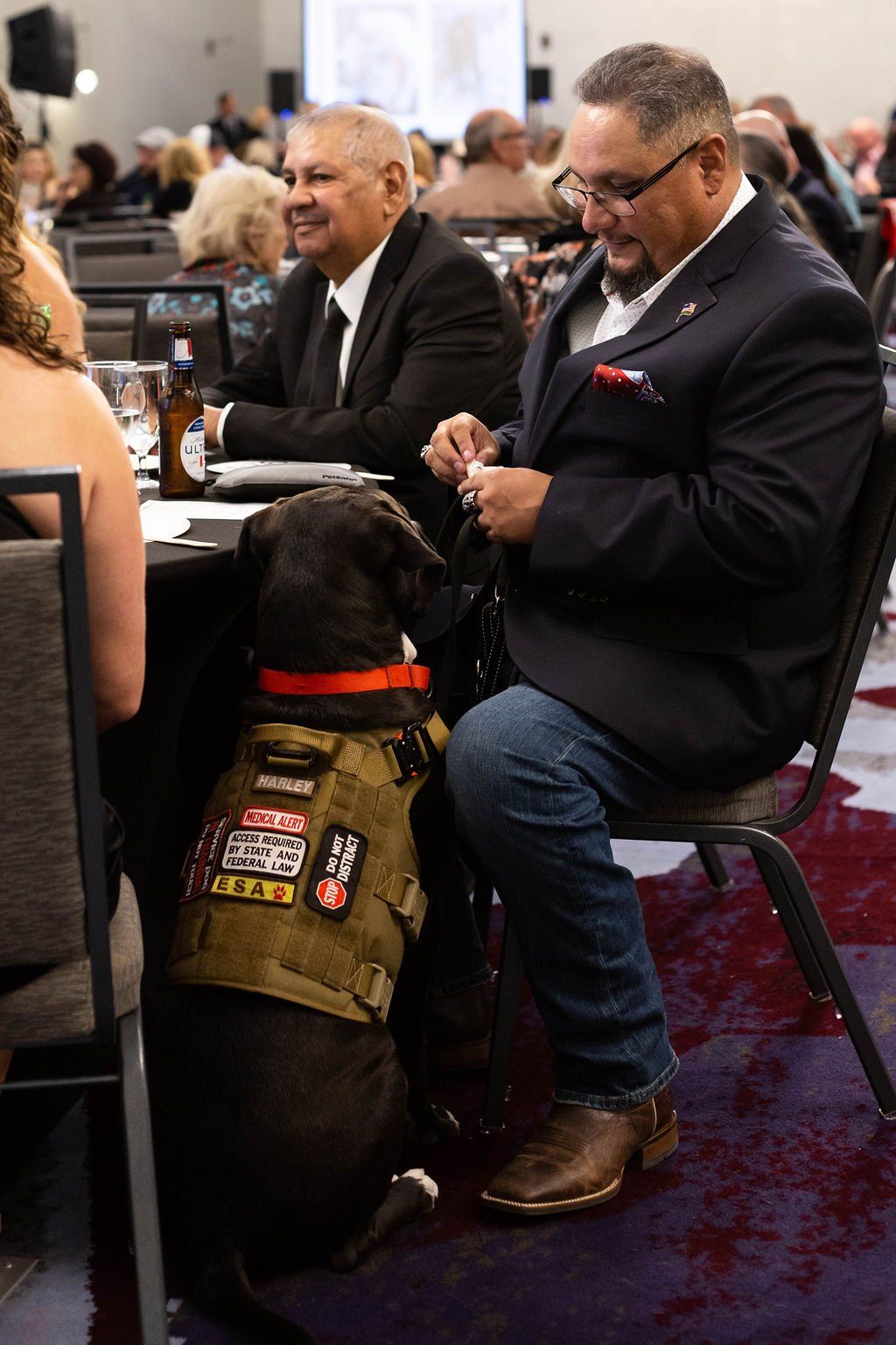 A service dog in a tactical vest sits beside a person at a table during an event in a banquet hall.