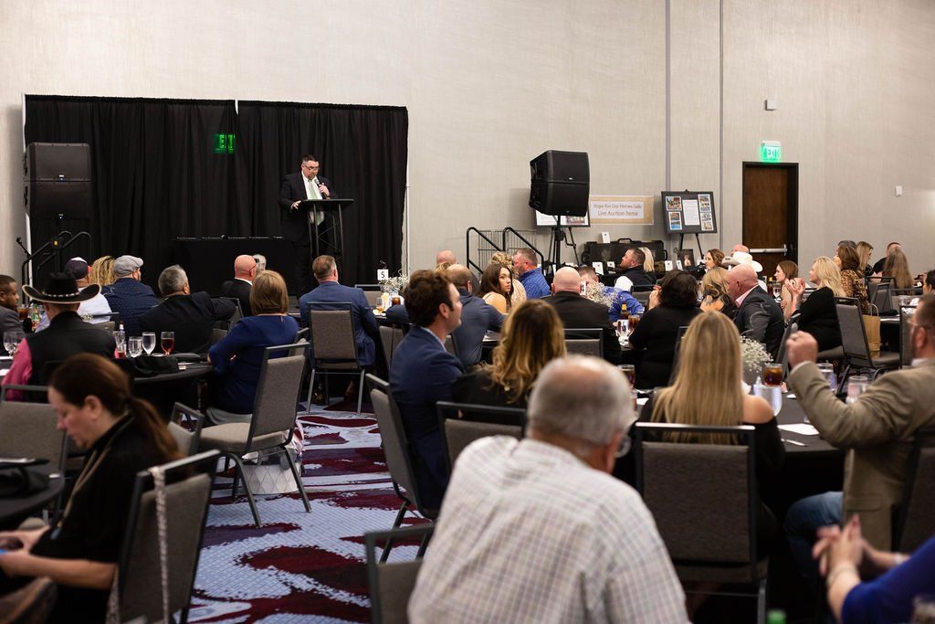 A speaker stands at a podium in a large, carpeted conference room addressing an audience seated at round tables.