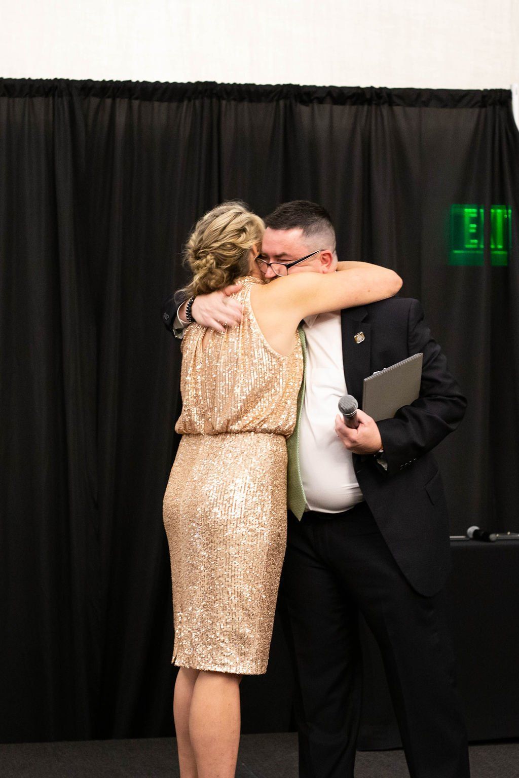 A woman in a gold sequined dress hugs a man in a black suit holding a microphone and a tablet against a black backdrop.