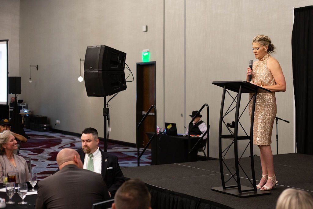 A person in a shimmering gold dress stands at a podium on a stage, speaking into a microphone before an audience.
