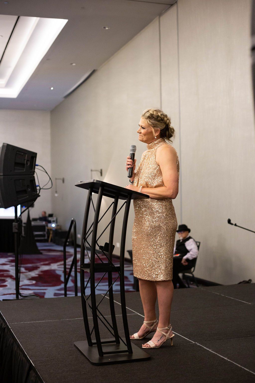 A person in a gold sequin dress speaks into a microphone at a podium on a stage in a large, modern conference room.