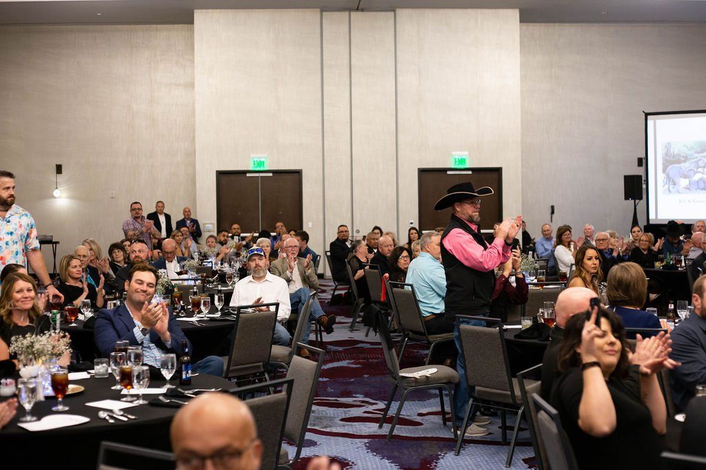 People gathered in a hotel ballroom at tables, some clapping, with a person in a cowboy hat standing and gesturing.