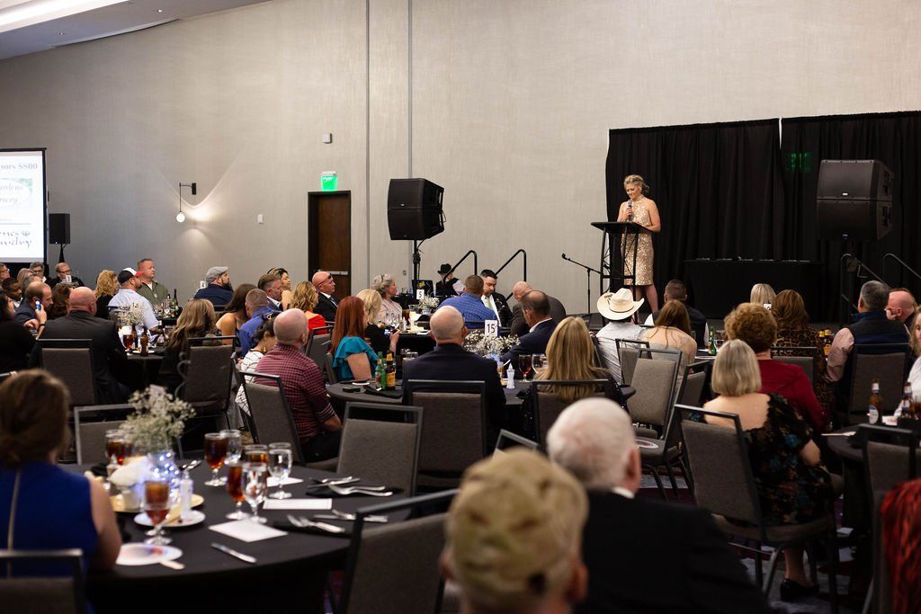 A speaker stands at a podium at the front of a crowded banquet hall, addressing attendees seated at round tables.