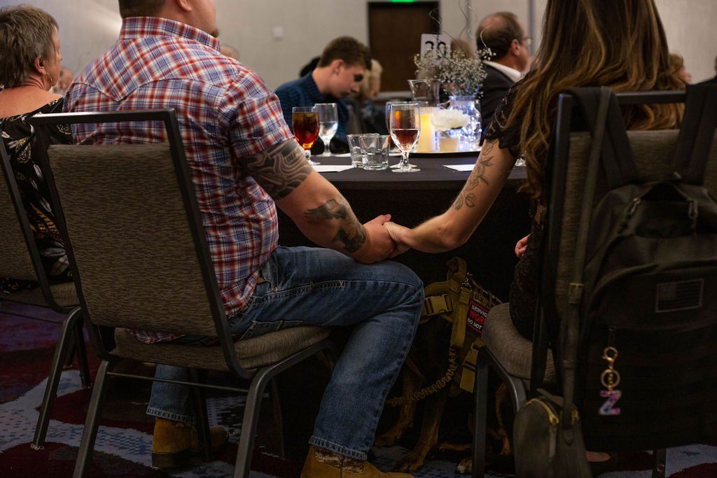 A couple sits at a round table in a dim room, holding hands across the table while seated in chairs.