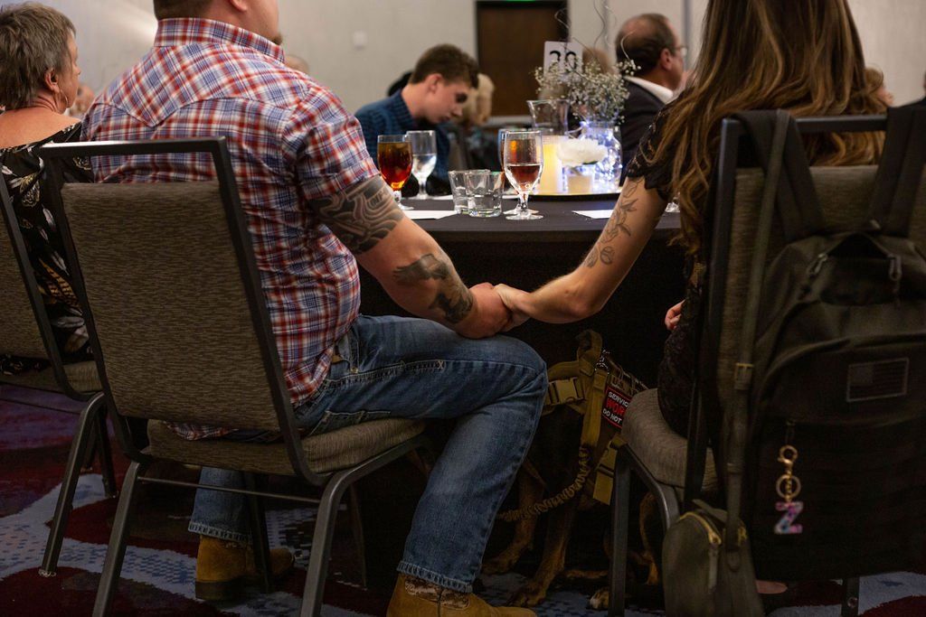 A couple sits at a table in a dimly lit room, holding hands across the table while others sit nearby.