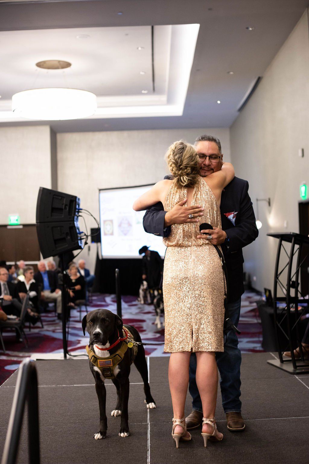 A woman in a gold dress hugs a man in a blazer on a stage, with a service dog wearing a vest standing in the foreground.