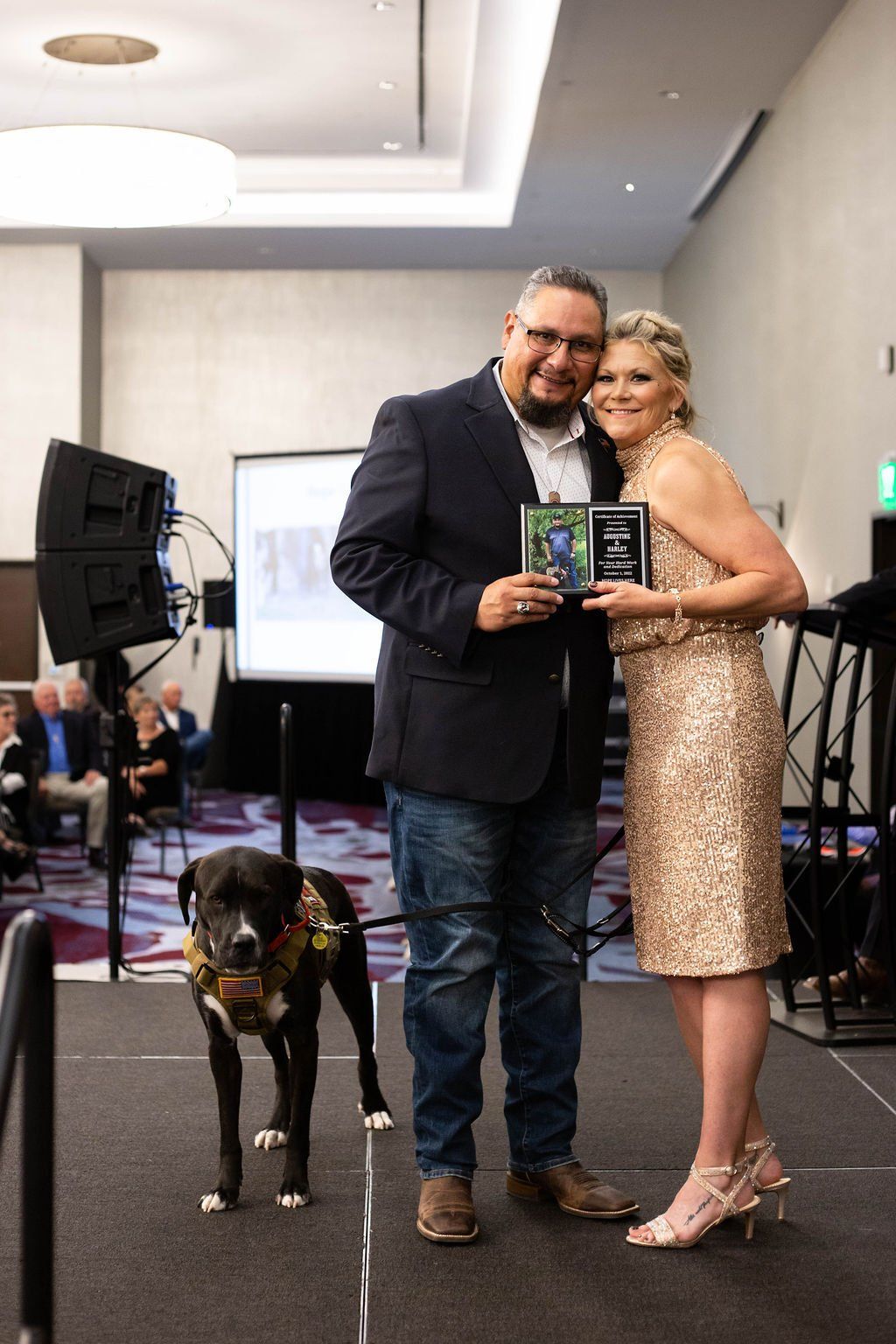 A person in a suit and a person in a sequined dress hold awards on stage next to a dog wearing a service harness.