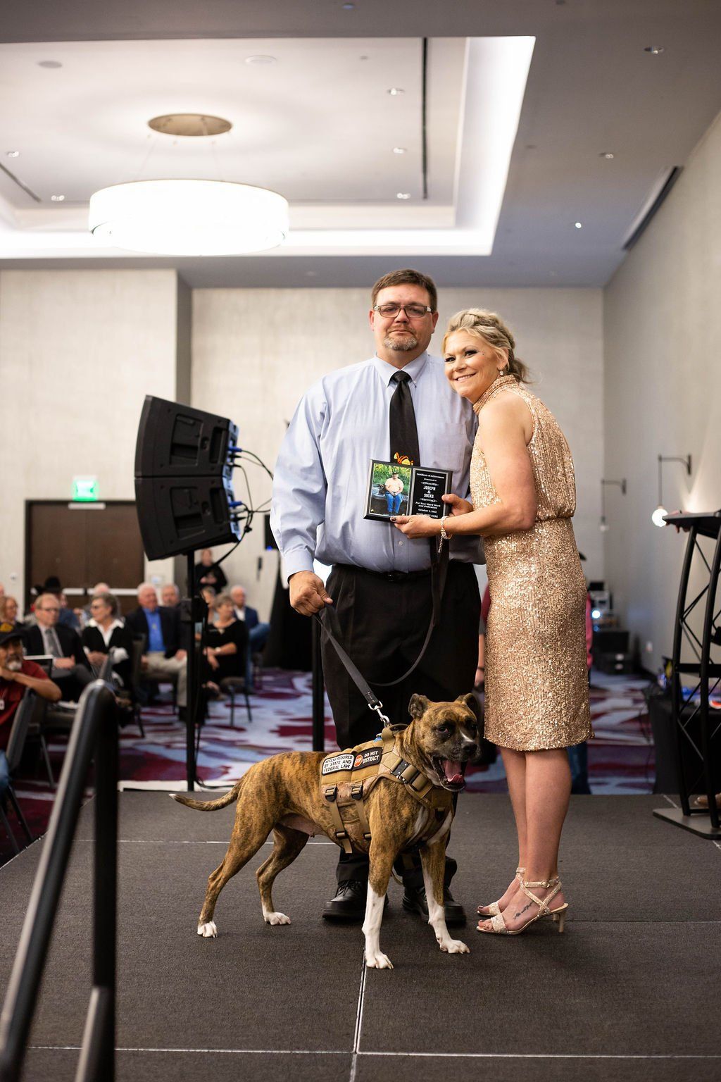 A person and a partner stand on a stage with a brindle service dog, holding an award in an event hall.