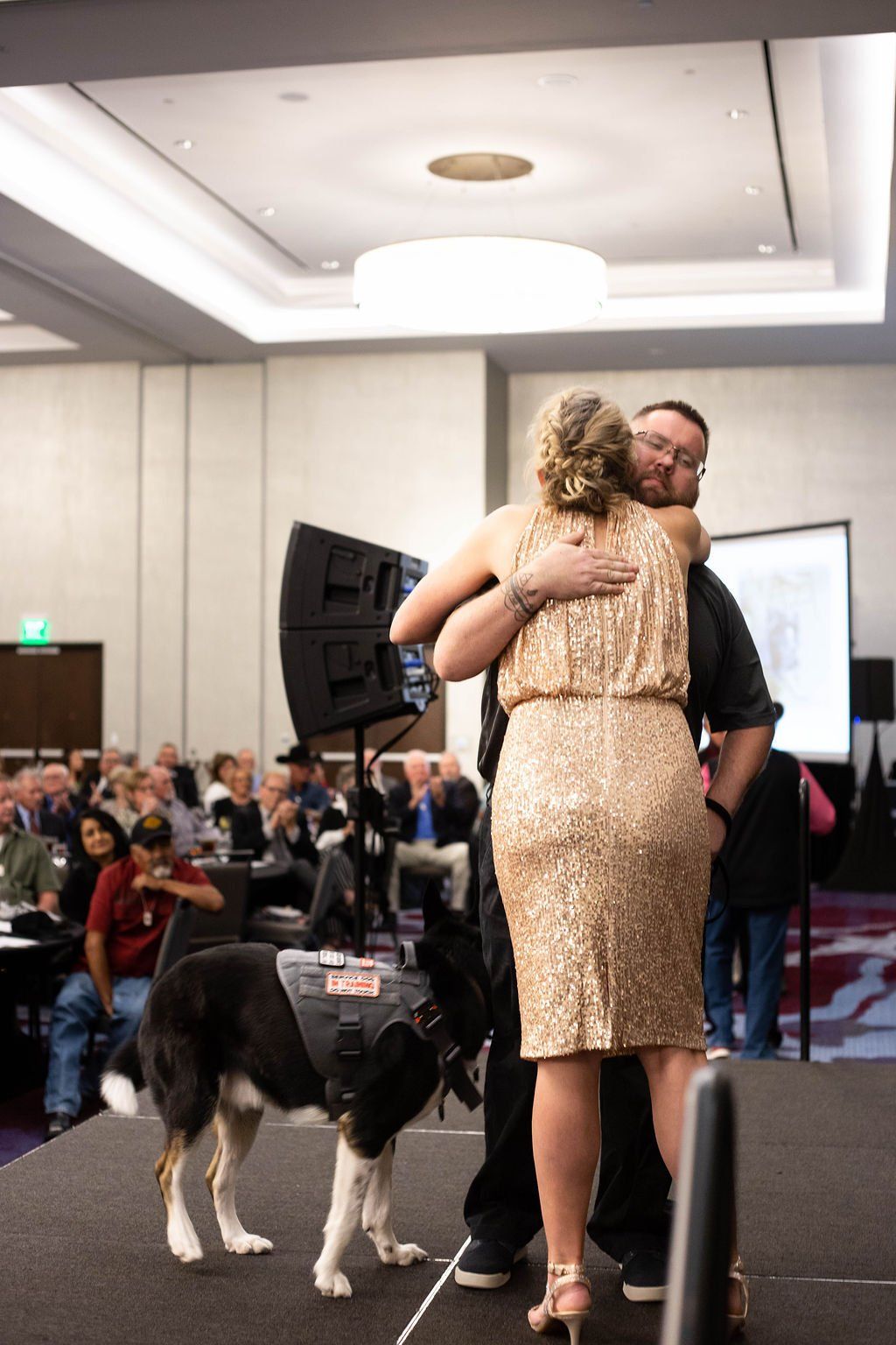 A woman in a gold sequined dress hugs a man on a stage, while a service dog in a harness stands beside them.