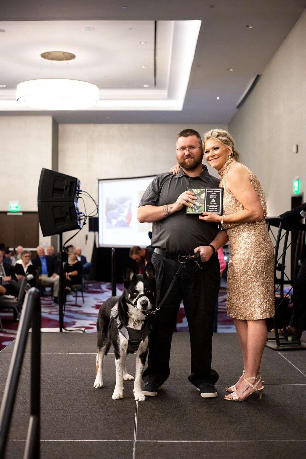 A man and woman stand together on a stage holding an award, with a service dog beside them in a conference room.