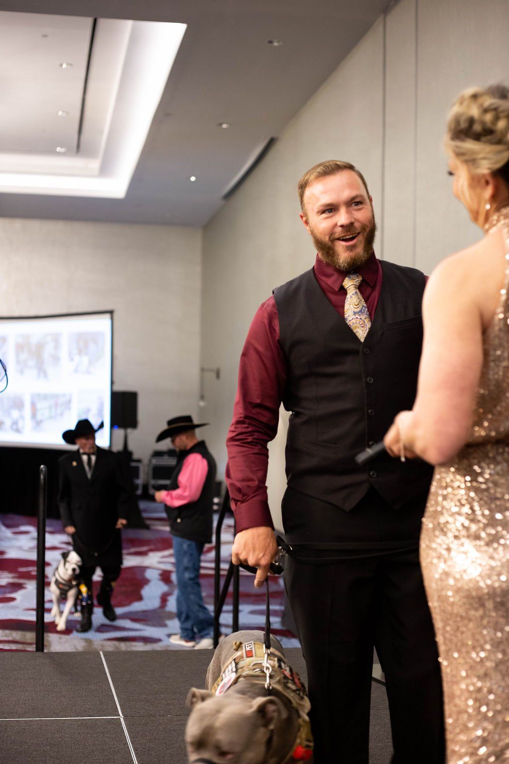 A man in a maroon shirt and vest stands on a stage with a dog, facing someone in a sequined gown at an indoor event.
