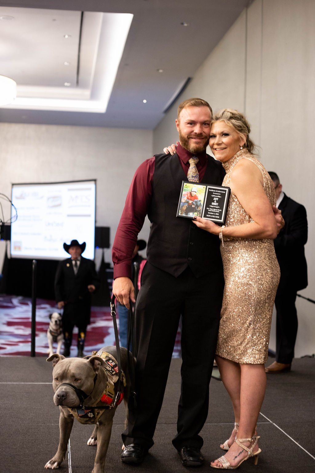 A man and a woman in formal attire stand on a stage holding a plaque, accompanied by a service dog on a leash.