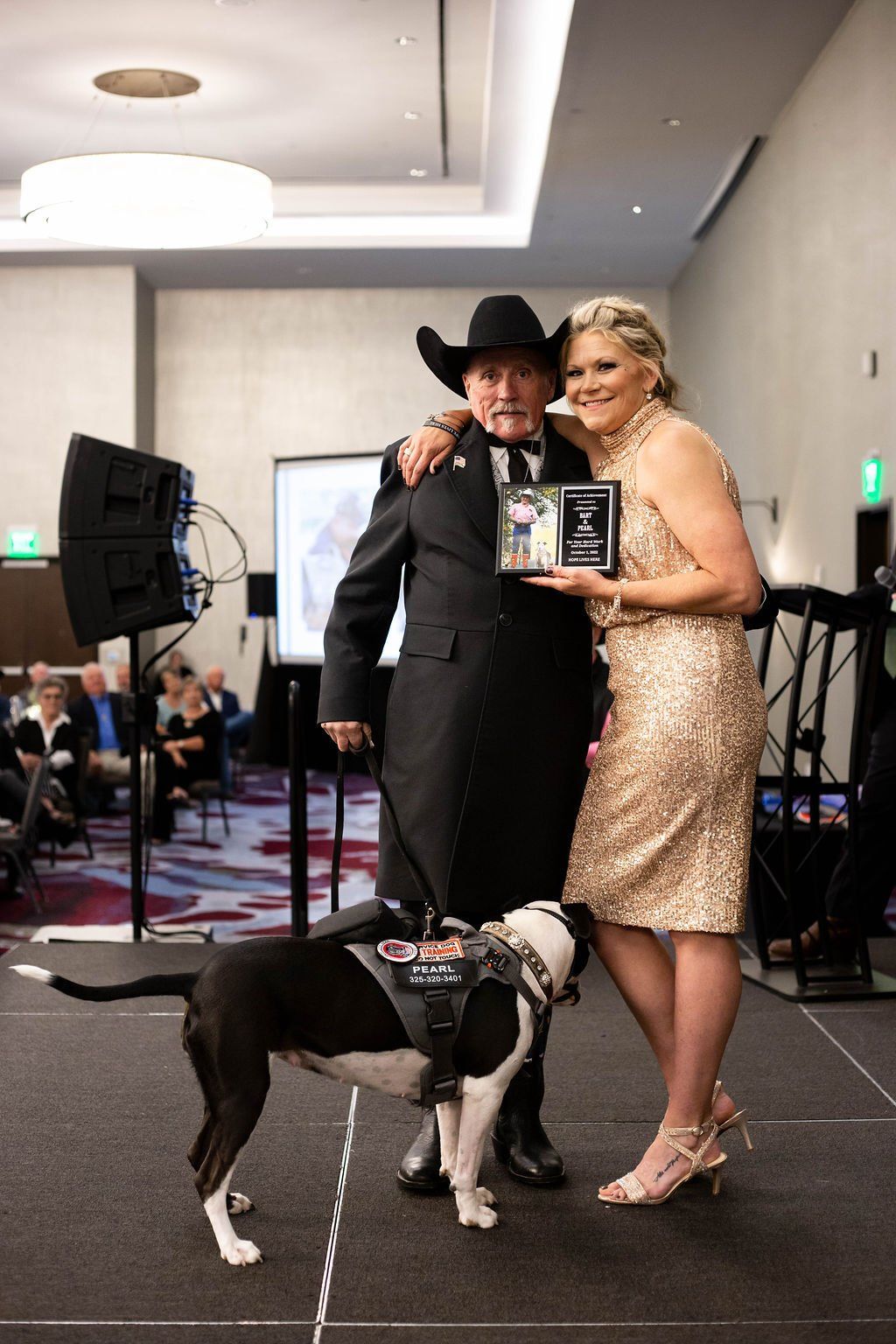 A person in a suit and cowboy hat and a person in a sequined dress stand with an award, accompanied by a service dog.