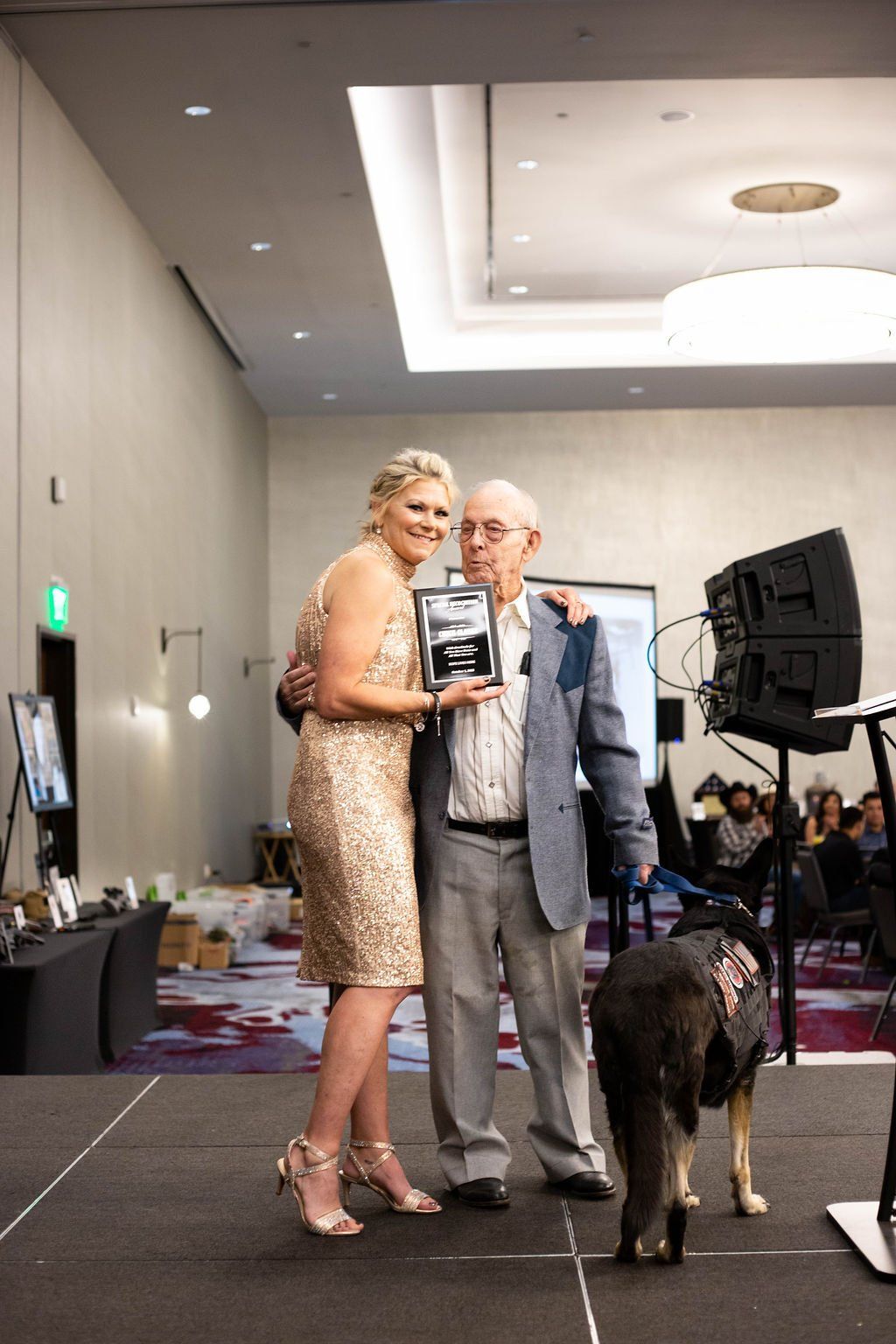 A person in a gold dress and a person in a blazer stand on a stage holding a plaque with a service dog standing nearby.