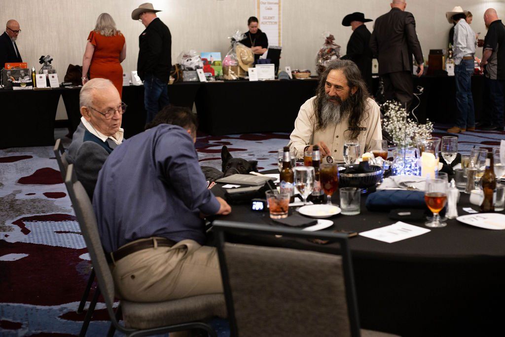 People at a formal event, with two individuals seated at a round table in the foreground and others standing near booths.