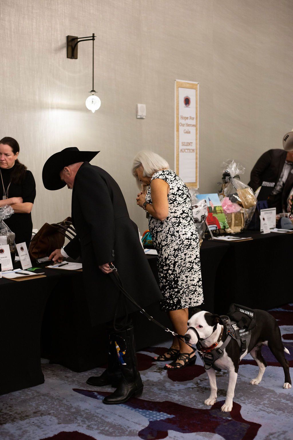 A person in a black coat and cowboy hat stands at a table with a service dog, while another person looks at items nearby.