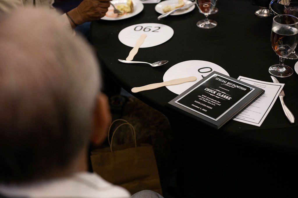A view over a shoulder at a table with white signs labeled 062, silverware, drinks, and a small event program.