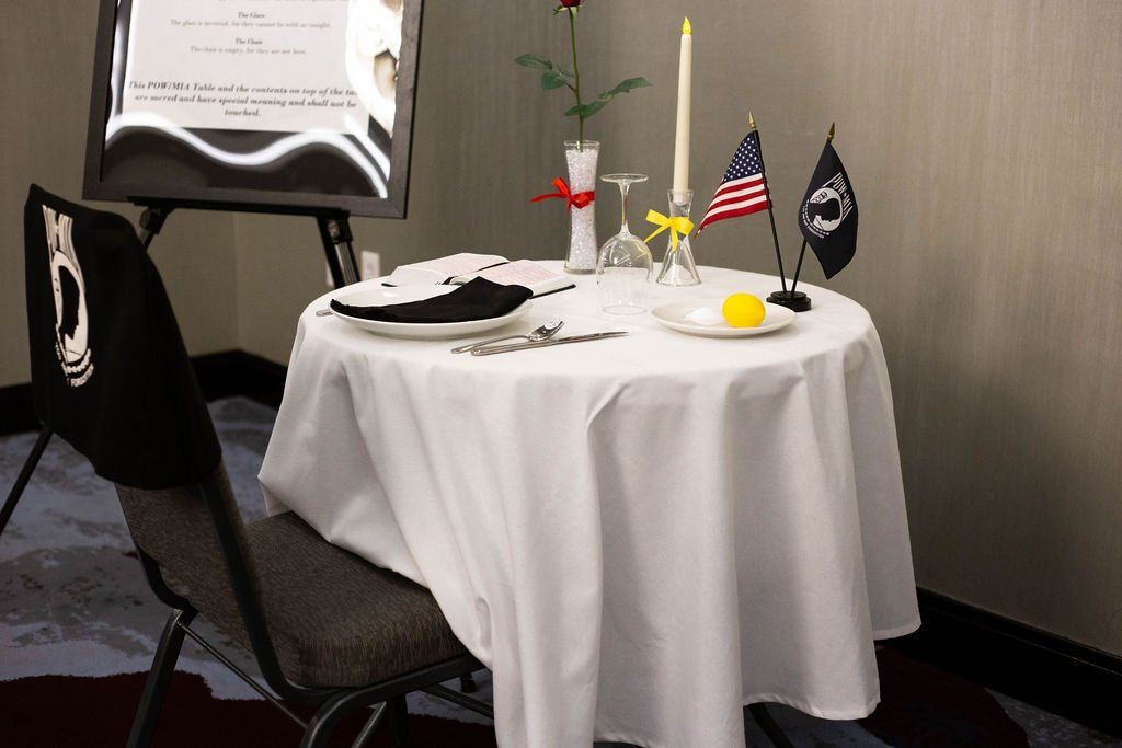 A POW/MIA remembrance table with a single place setting, a candle, a rose, and US and POW/MIA flags on a white tablecloth.