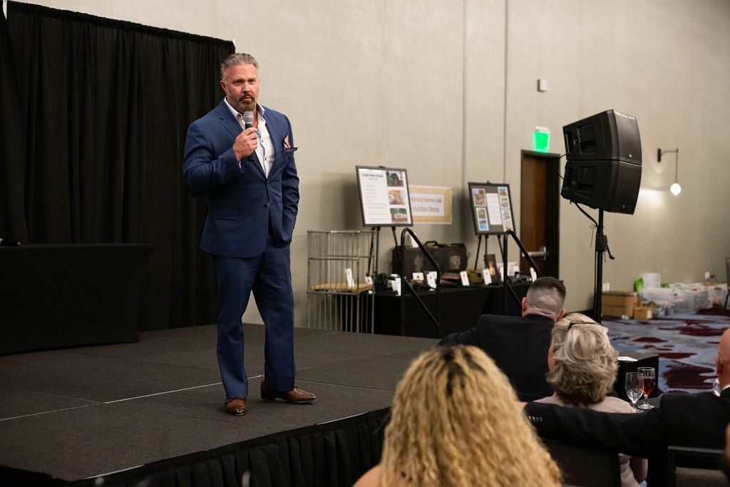 A person in a blue suit speaks into a microphone on a stage before an audience in a banquet hall.