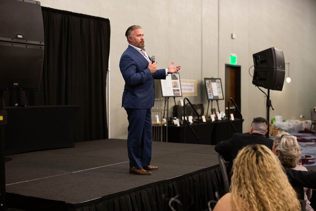 A man in a blue suit speaks into a microphone on a stage before an audience in a conference room.