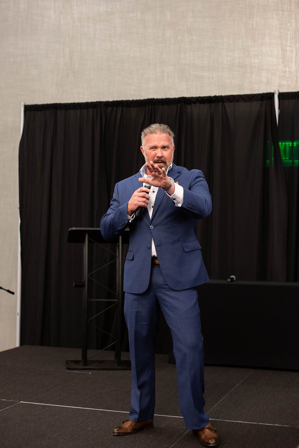 A person in a blue suit stands on a stage with a microphone, gesturing with one hand in front of a black curtain.